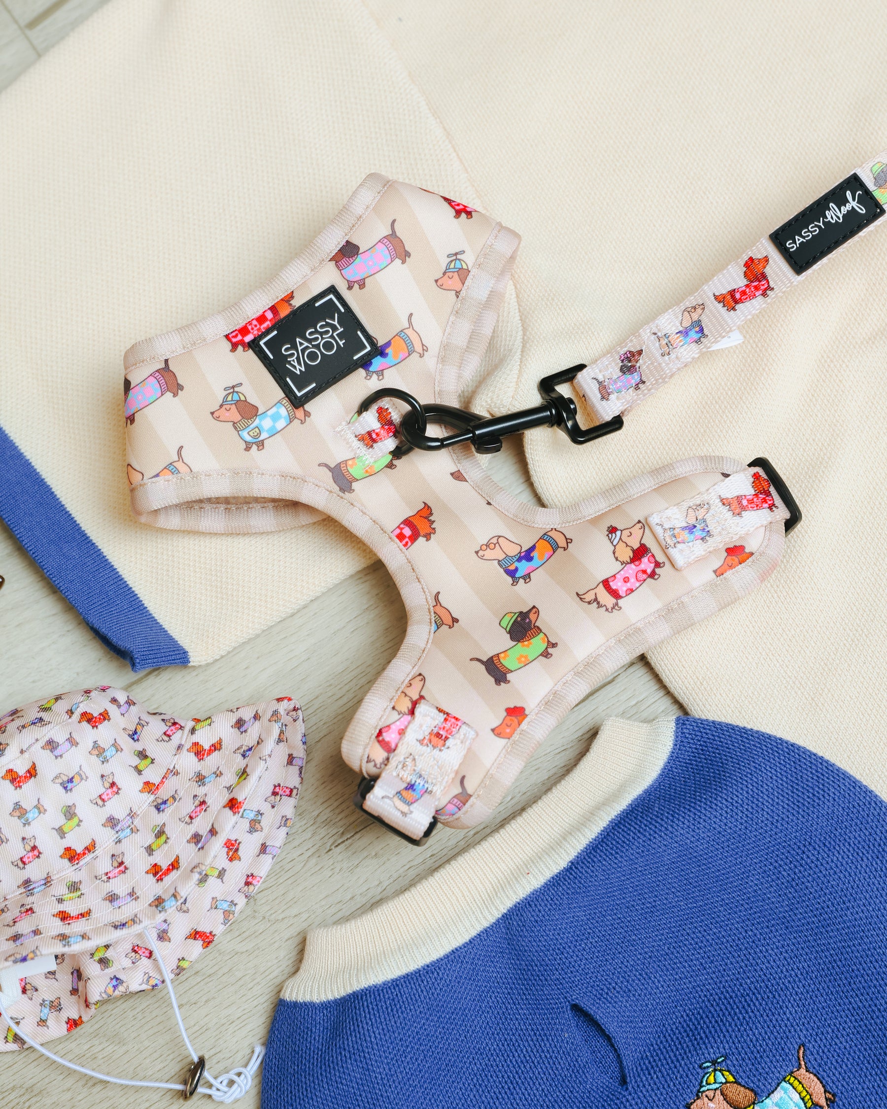 The Sassy Woof Dog Two Piece Bundle - Sunbean, an adjustable beige harness and leash set adorned with colorful cartoon Doxie prints, is shown displayed on folded clothes and a hat featuring a matching dog design.