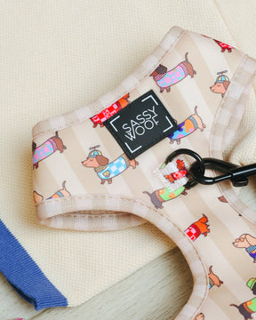 The Sassy Woof Dog Adjustable Harness - Sunbean, featuring a playful print of cartoon dogs in sweaters and a beige background, is adorned with the signature Sassy Woof label and includes a sturdy black leash clip. Resting on a soft, light fabric surface next to blue-edged material, this no-pull harness combines style and practicality—ideal for dachshunds or other small pups.