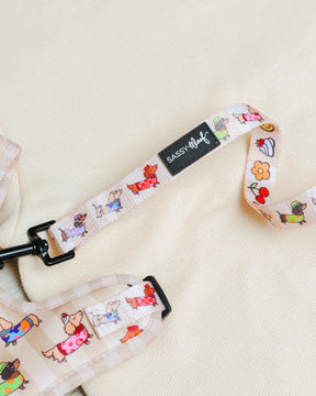 A close-up of the "Dog Leash - Sunbean" by Sassy Woof, showcasing its playful cartoon animal and fruit pattern with a prominent black Sassy Woof label, lies on a light cream fabric background—making it an ideal pick for anyone looking for coordinating dog accessories.