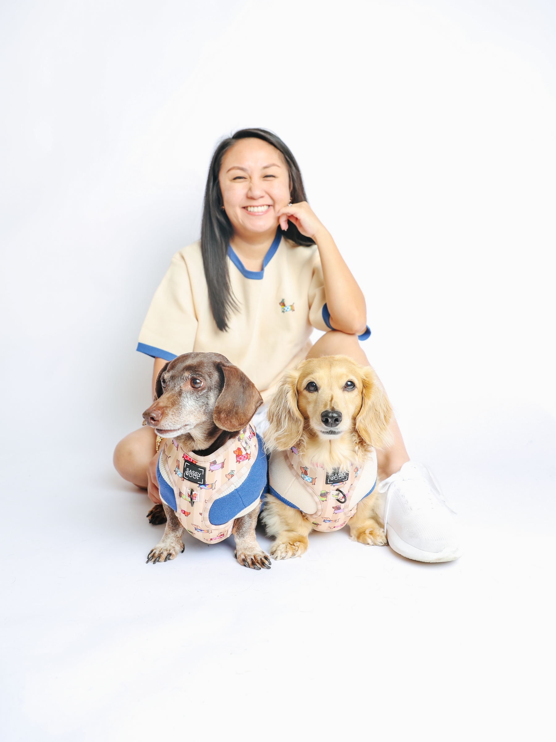 A smiling woman sits on the floor with two dachshunds in matching Sassy Woof Dog Adjustable Harnesses - Sunbean in front of her, against a plain white background.