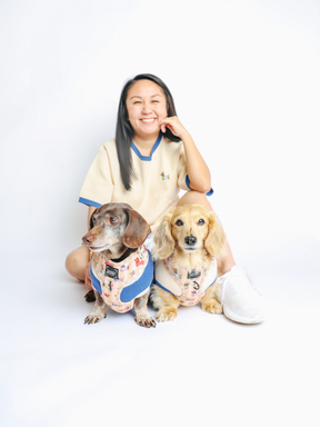 A smiling woman sits on the floor with two dachshunds in matching Sassy Woof Dog Adjustable Harnesses - Sunbean in front of her, against a plain white background.