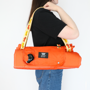 A person with long brown hair carries the Sassy Woof Dog Picnic Blanket - Hot Dawg, featuring an orange design inspired by dachshunds, a yellow strap with dog illustrations, a black label, and a built-in dispenser for waste bags.