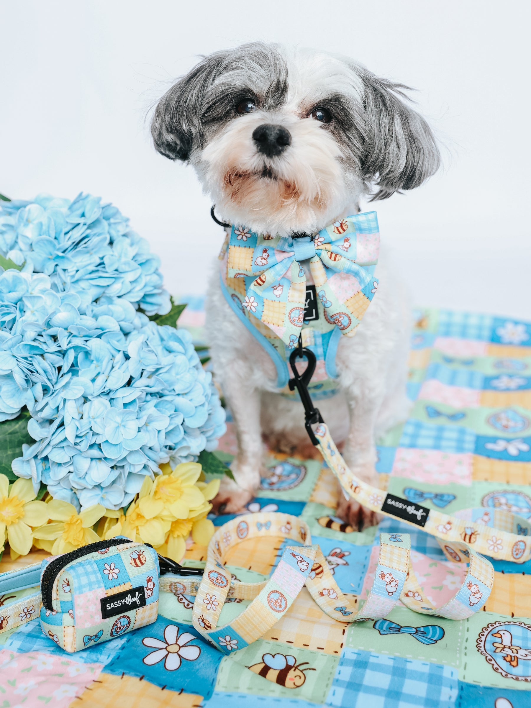 A small gray and white dog sits on a quilted blanket next to blue and yellow flowers, wearing Sassy Woof's Patch Perfect adjustable harness and leash. A matching accessory pouch is placed in front of the dog.
