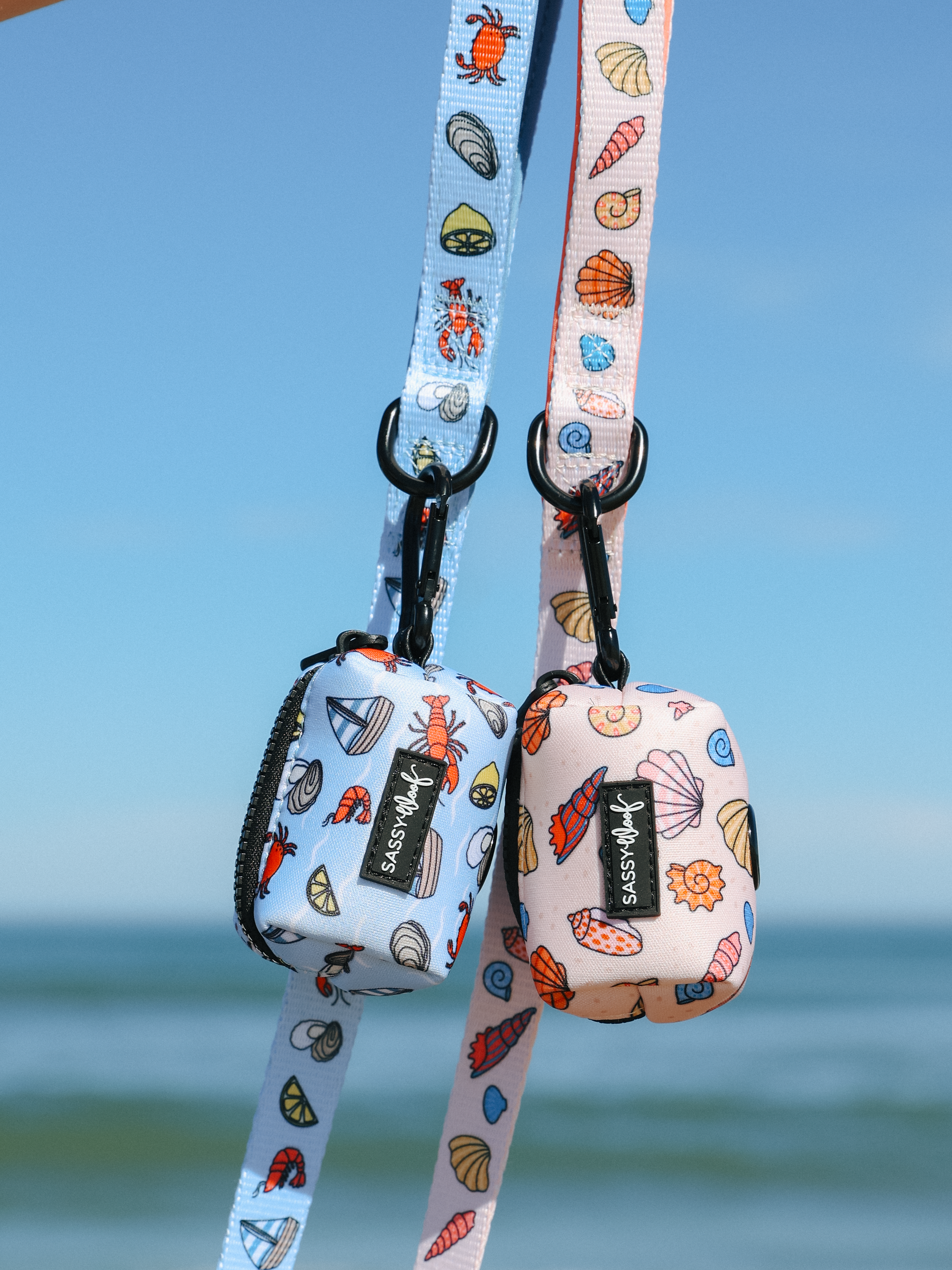 Two small pouches with seashell and crab patterns hang from matching lanyards against a blue sky and ocean. One pouch is blue, the other pink, both labeled “Waggyss.” Shown with the Sassy Woof Dog Leash - Shell Yeah, perfect for beachy adventures.