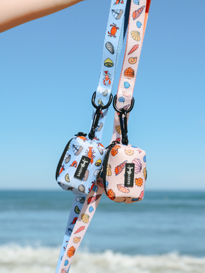 A Sassy Woof Dog Leash from the Coastal Cuties collection, featuring blue and pink seashell and sea creature designs, is displayed with a matching lanyard against a bright sky and ocean backdrop.