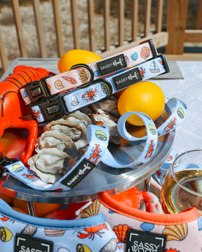 The Sassy Woof Dog Collar - Shell Yeah features colorful, ocean-inspired patterns. It’s displayed with matching leashes, lemons, oysters, a lobster toy, white wine, and pet bandanas for a lively seaside vibe.