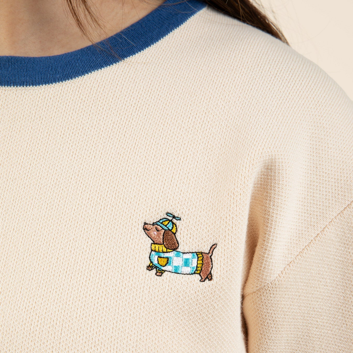 A close-up of the cream-colored Sassy Woof x Sunbean Human Tee by Sunbean, accented with blue trim and featuring an embroidered dachshund sporting a blue hat and a blue-and-yellow checkered sweater—perfect for those who love the matching dog and owner tee trend.