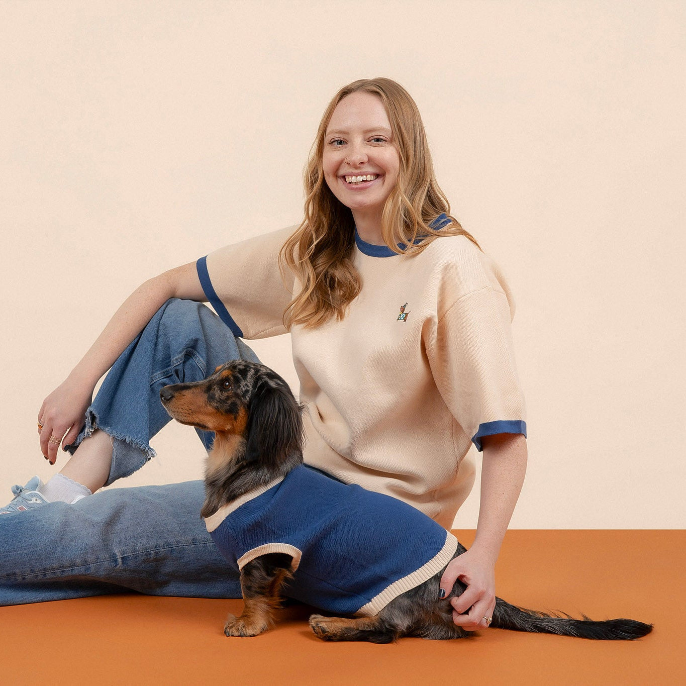 A smiling woman in a beige and blue shirt sits on the floor next to a small dachshund wearing the Sassy Woof x Sunbean Dog Tee by Sunbean. Both face the camera against a plain, light background, perfectly showcasing their matching dog and owner looks from the collaborative collection.