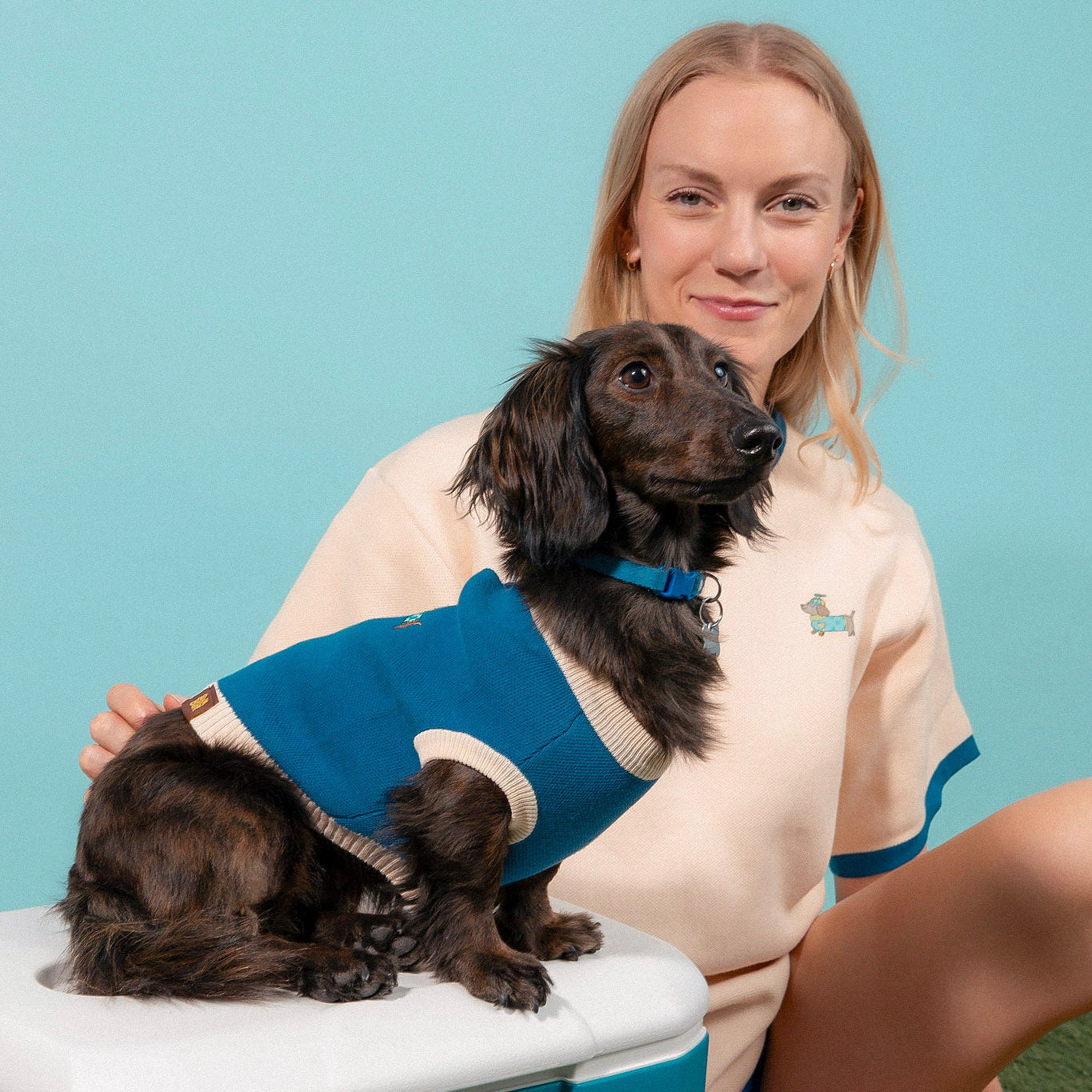 A woman wearing a cream Sunbean Sassy Woof x Sunbean Dog Tee sits beside a long-haired dachshund in a matching blue sweater, both sporting coordinating dog and owner outfits against a light blue background.