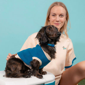 A woman wearing a light-colored shirt sits and smiles while gently holding a dachshund dressed in the Sunbean Sassy Woof x Sunbean Dog Tee. The background is a solid light blue color.