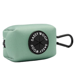 The Sassy Woof Dog Waste Bag Holder - Sage is a light green, rectangular neoprene bag holder with a black circular logo and a black clip for easy attachment to your dog’s leash, harness, or collar.