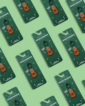 Sassy Woof Dog Collar Tags—Hug Me Bear charms—are arranged in a diagonal pattern on a green background, highlighting the colorful pet tags and playful designs.