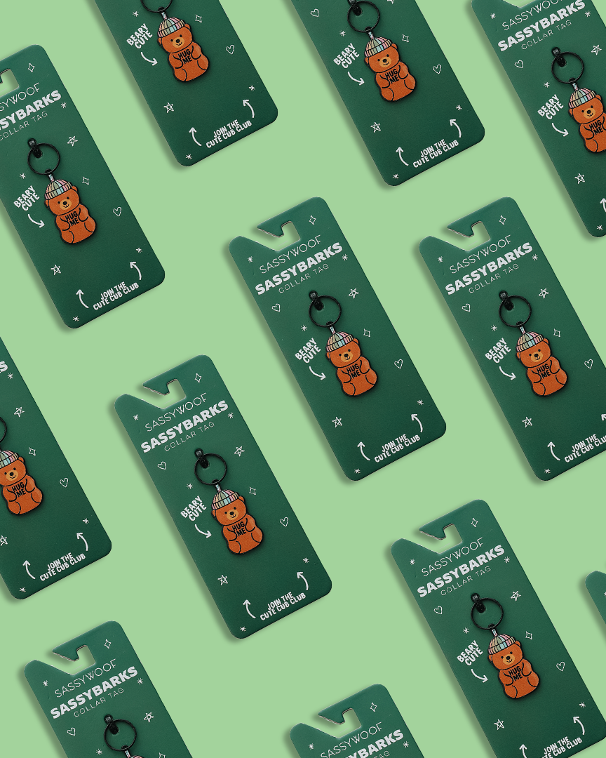 Sassy Woof Dog Collar Tags—Hug Me Bear charms—are arranged in a diagonal pattern on a green background, highlighting the colorful pet tags and playful designs.