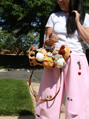 A woman in a white tee and pink pants stands outside, smiling while holding a Sassy Woof Blind Box handbag adorned with collectible stuffed toys and a dachshund plush keychain. Trees and grass are visible in the background.