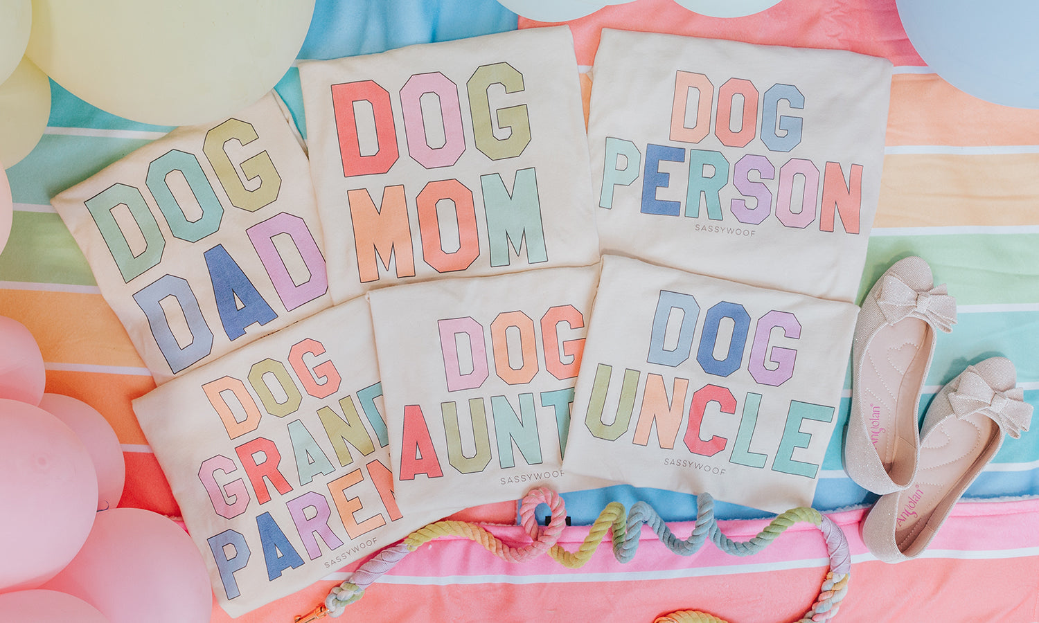 Six pastel-colored shirts with phrases: Dog Dad, Dog Mom, Dog Person, Dog Grandparent, Dog Aunt, and Dog Uncle, laid out on a striped blanket with balloons, rope toy, and shoes nearby.