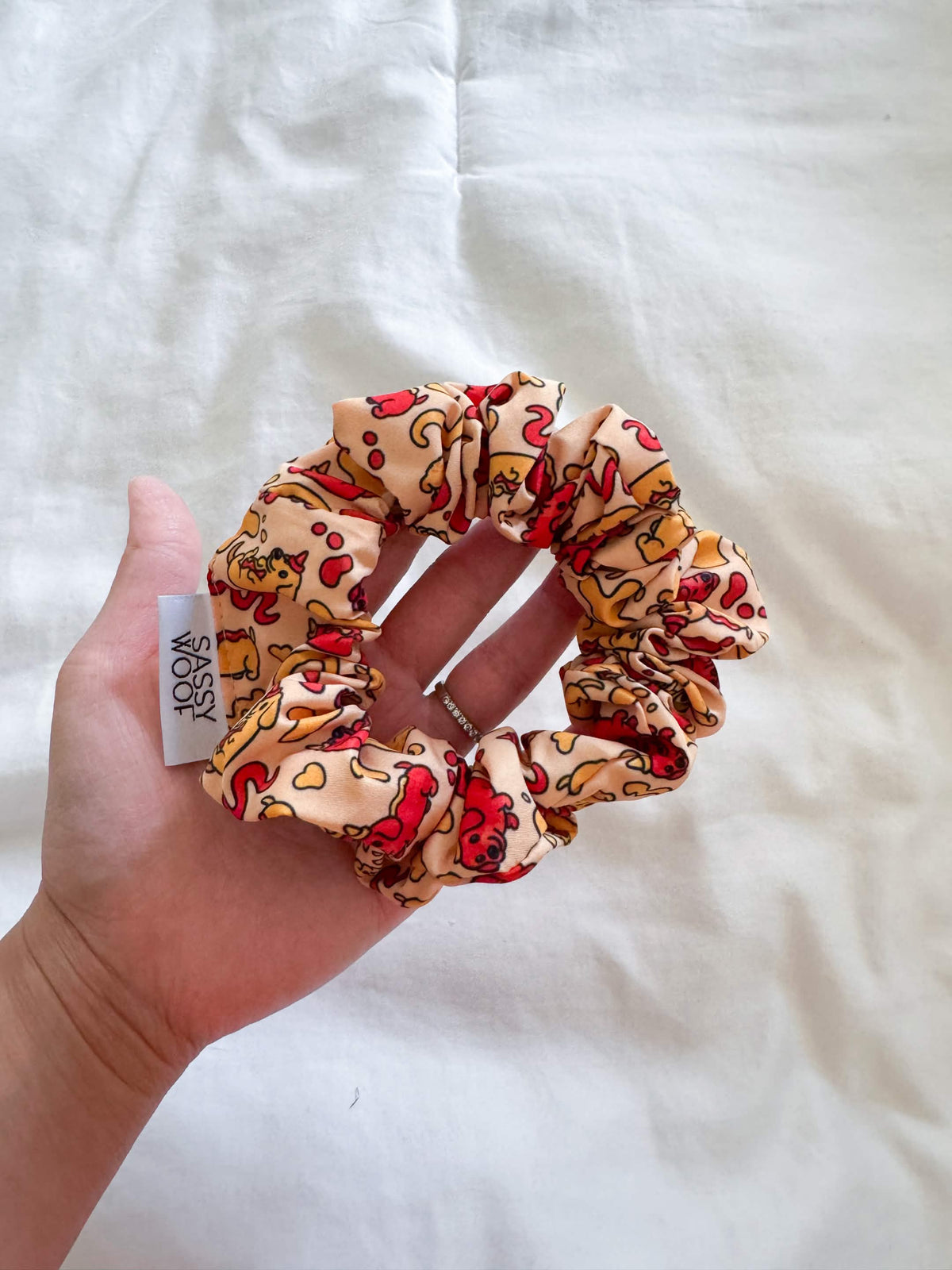 A hand holds the Sassy Woof "Hot Dawg" scrunchie, featuring a colorful pizza slice print from their dachshund-inspired collection. The scrunchie has a small white Sassy Woof tag and is placed on a white blanket.