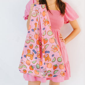 A woman in a pink dress holds the Sassy Woof Fruity Reusable Bag, a pink foldable shopping bag featuring colorful cat, dog, fruit, and vegetable illustrations, against a plain white background.