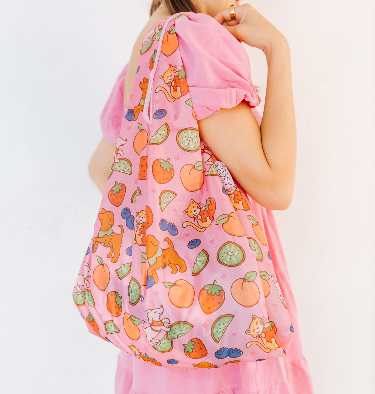 A person in a pink dress holds the Sassy Woof Fruity Reusable Bag, which features playful cat illustrations, fruit slices, and berries, set against a plain white background.