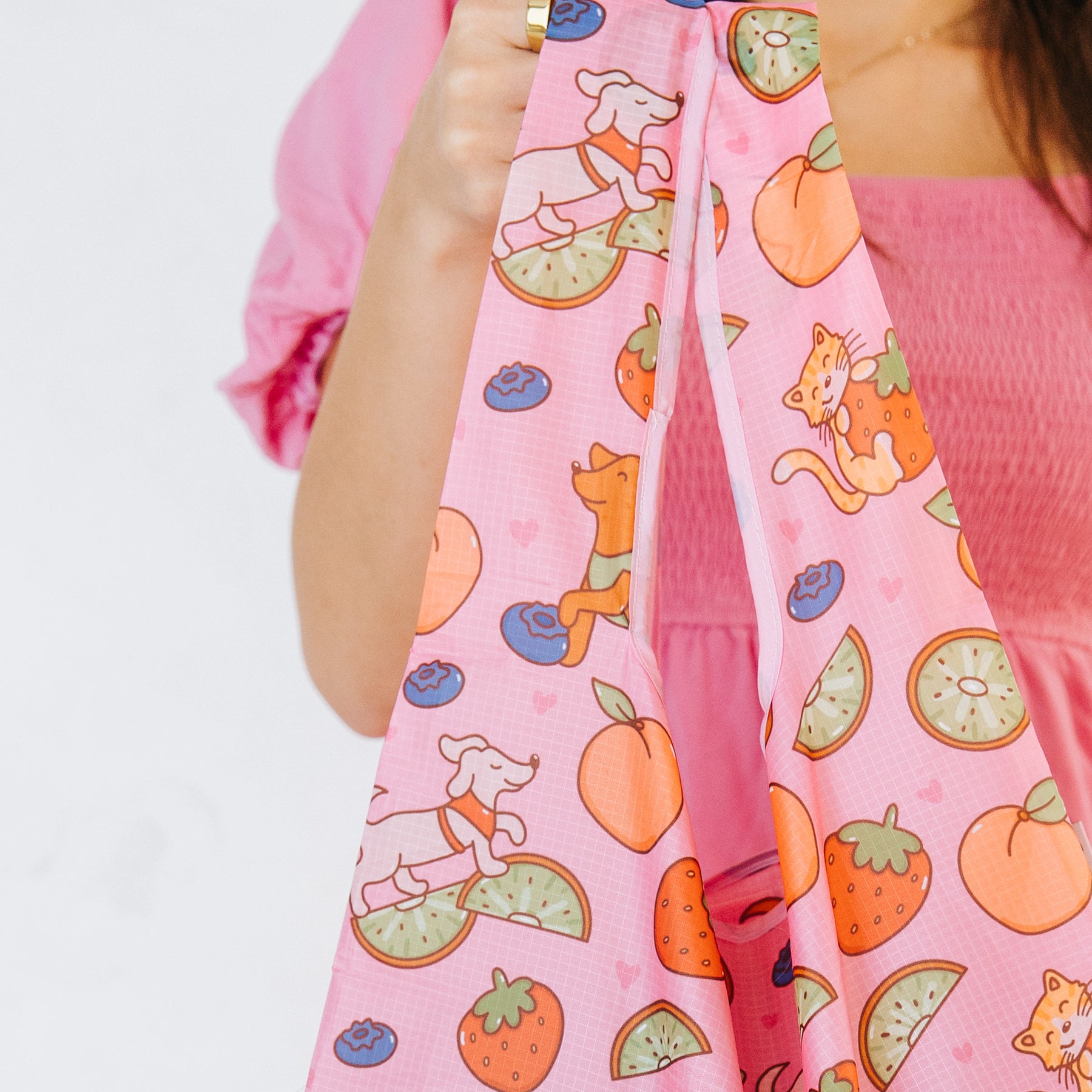 A person in a pink top holds the Sassy Woof Fruity Reusable Bag, featuring playful dog, cat, fruit, and heart designs.