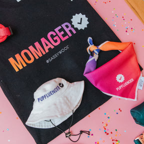 Black tote bag with MOMAGER text, a white hat labeled Pupfluencer, and the Sassy Woof Dog Bandana - Pupfluencer, a colorful lightweight cotton bandana, on a pink surface with confetti.