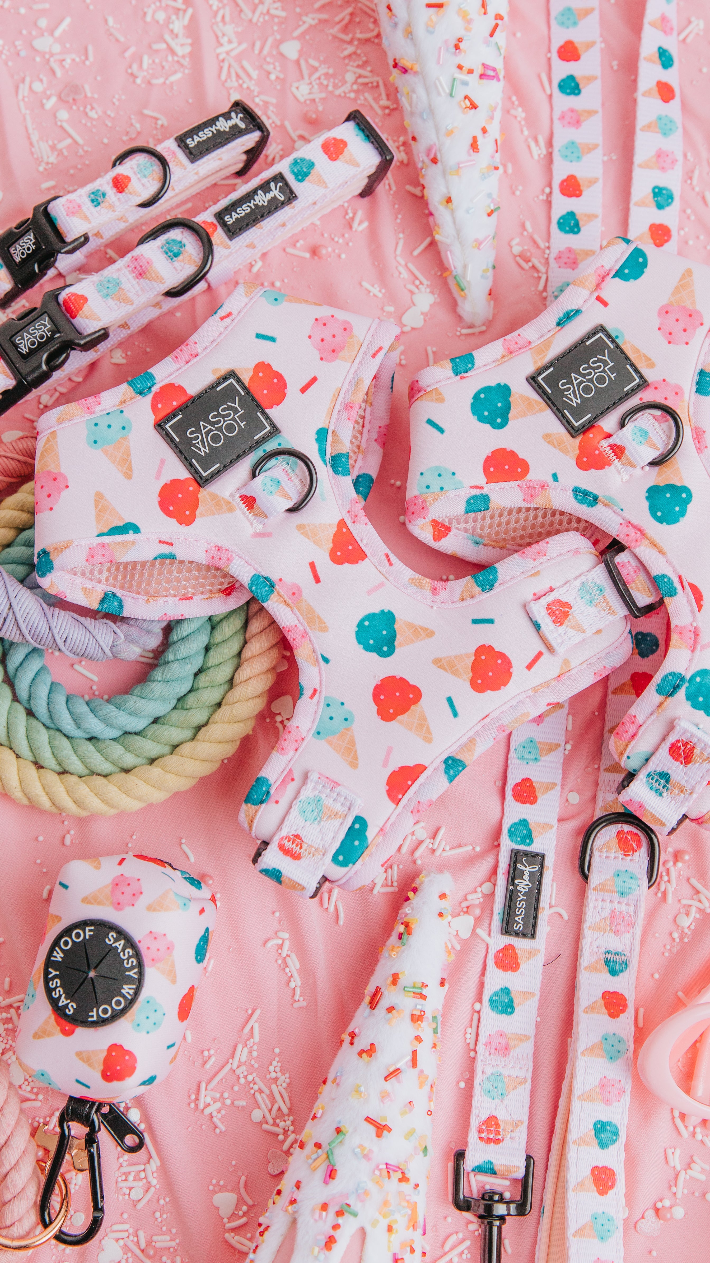 A flat lay of pink dog accessories, including harnesses, leashes, and collars, all decorated with colorful ice cream and popsicle patterns, displayed on a pink surface with sprinkled decorations.