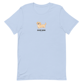 The Doxie Mom Tee by Sassy Woof is a light blue unisex shirt with a small tan dachshund illustration above bold black "DOXIE MOM" lettering on the chest.