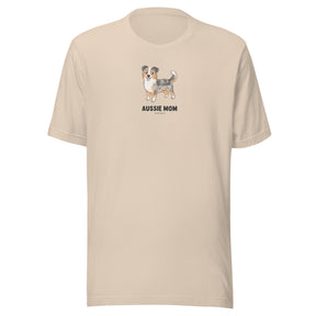 The Sassy Woof Aussie Mom Tee is a beige unisex cotton t-shirt with an Australian Shepherd illustration and bold "AUSSIE MOM" text on the chest—perfect for proud Aussie moms.