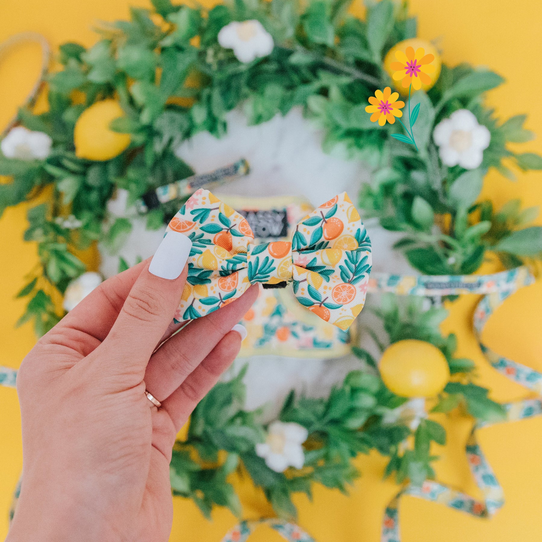 A hand with white nail polish holds the Sassy Woof Cat Bowtie - Zest Friends, featuring citrus and leaf patterns, in front of a lemon-adorned leafy wreath and white flowers on a bright yellow background.
