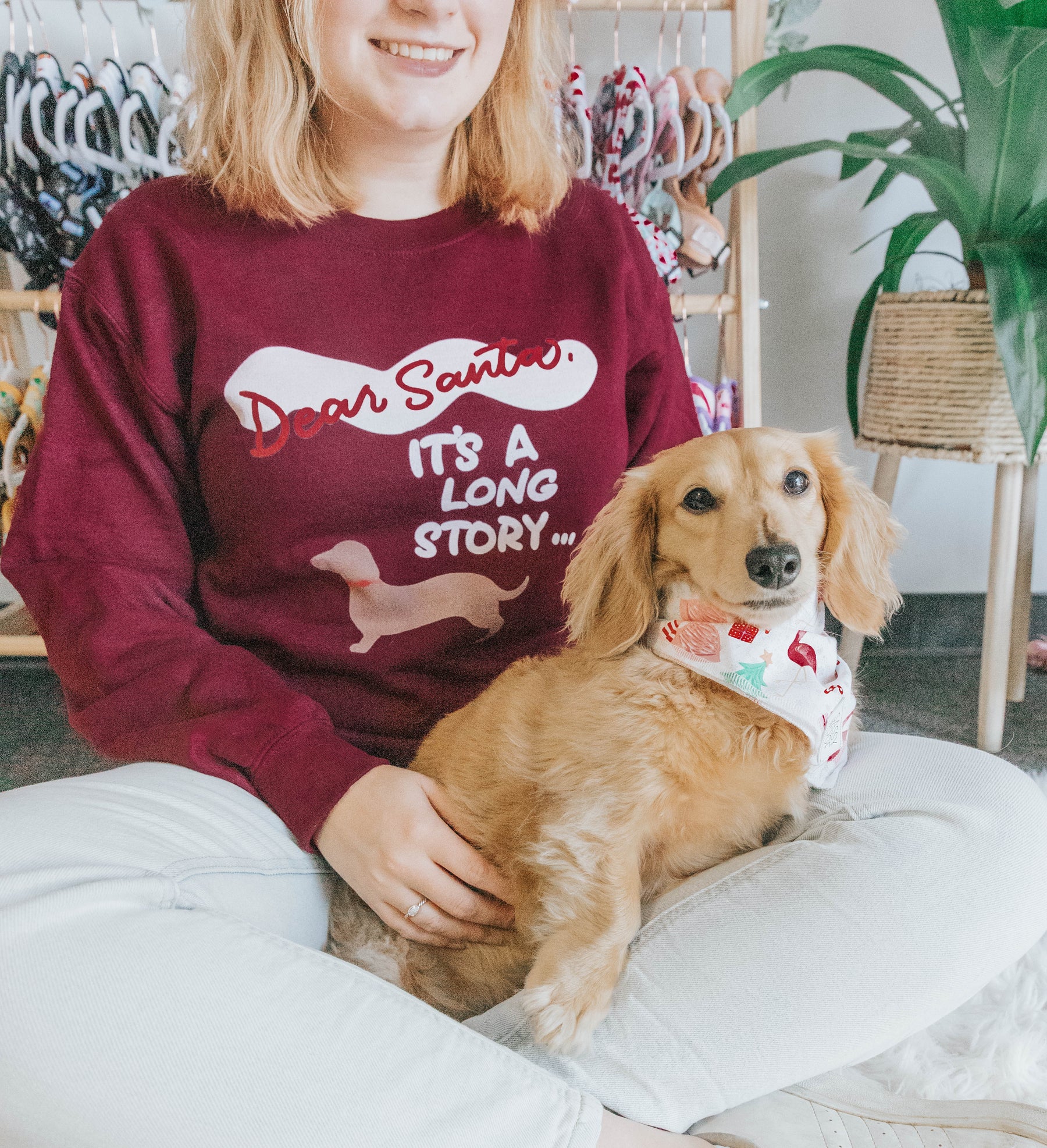 A woman sits cross-legged in a Sassy Woof Sweatshirt - Dear Santa, It’s a Long Story, holding a long-haired dachshund wearing a festive patterned bandana.