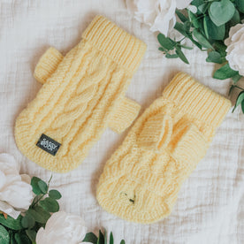 The yellow Sassy Woof Pet Cable Knit Sweater is shown on a white textured surface with green leaves and white flowers. Its cozy cable pattern matches fingerless mittens, and one features a small black Sassy Woof label.