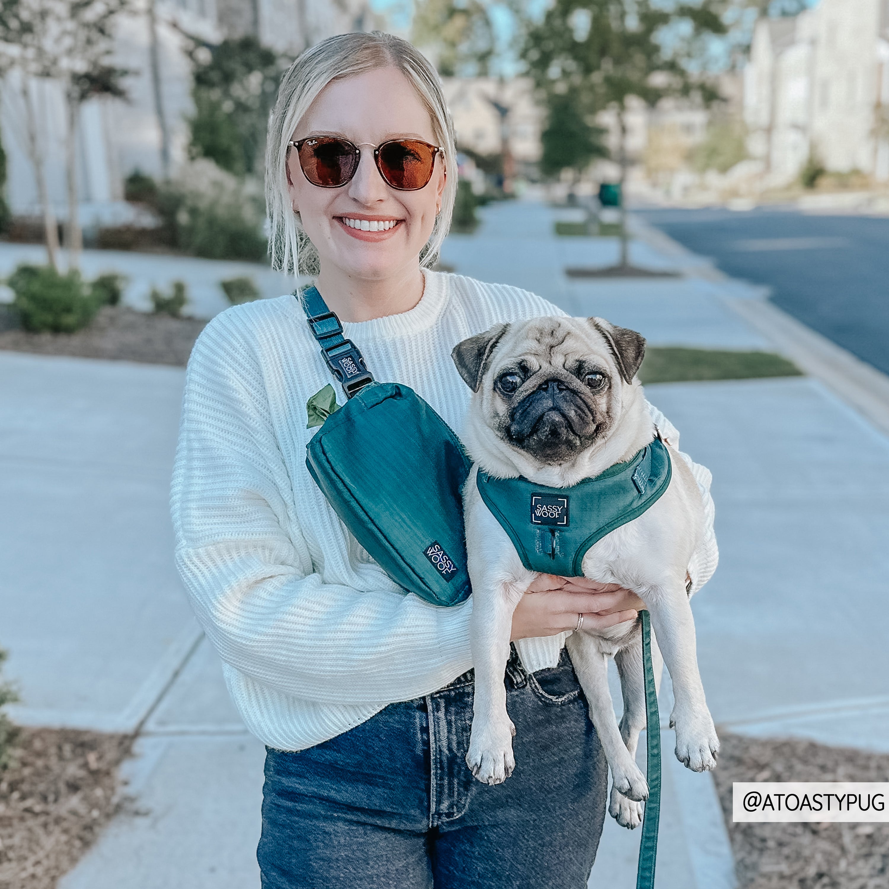 A woman in sunglasses and a white sweater smiles while holding a pug in a teal harness and matching Sassy Woof Forest Woof Pack (Bag Only) on a suburban sidewalk. Both sport coordinated accessories. Instagram: @ATOASTYPUG in the corner.