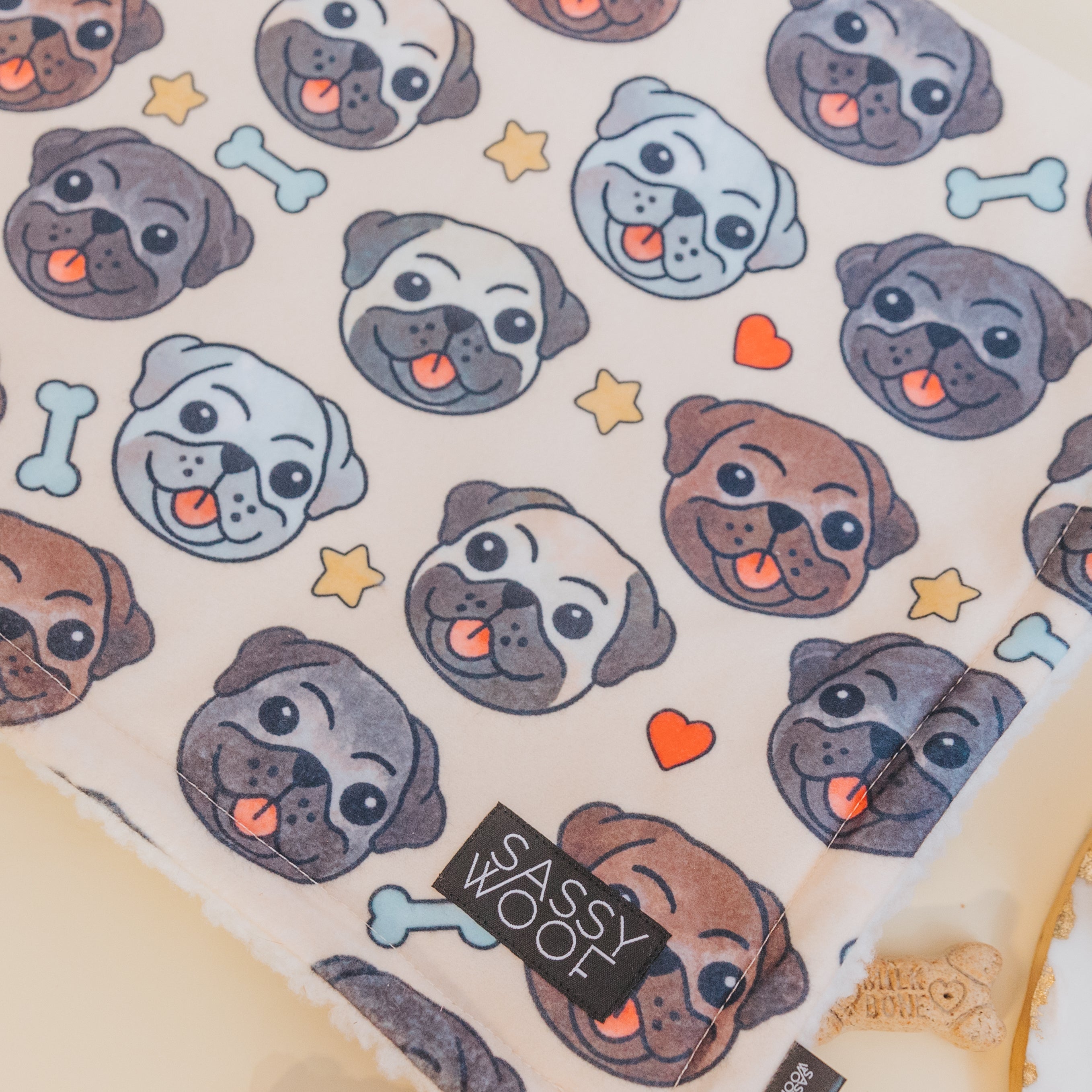 The Sassy Woof Pet Blanket - It's a Pug's Life is a soft microfiber blanket with colorful cartoon pug faces, bones, stars, and red hearts. A black “SASSY WOOF” label is sewn on the edge for an extra cute detail.